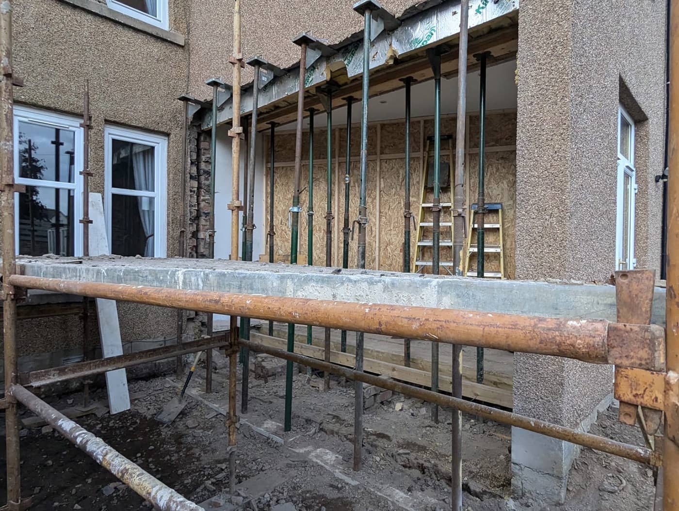 Adjustable steel props supporting the structure above a wide opening at the rear of a pebble-dashed house, with scaffolding in front